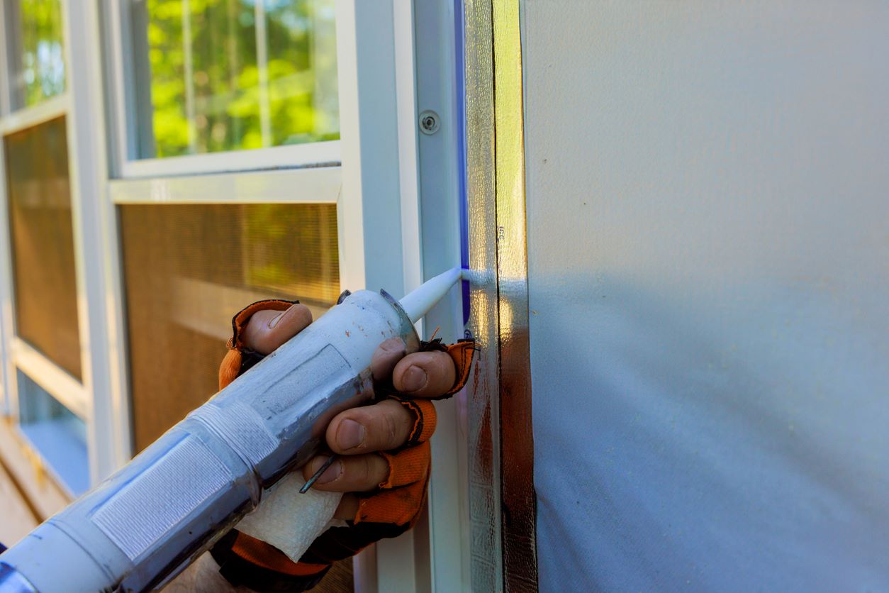 A person sealing the opening near the window with caulk to prevent spiders and other pests.